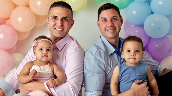 gay dads holding twin babies in front of pastel rainbow balloons