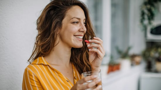 woman smiling wearing dark yellow button down shirt taking red vitamin holding clear short glass of water