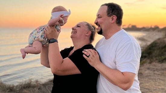 a new mother and father hold their baby girl up in the air against a beach sunset