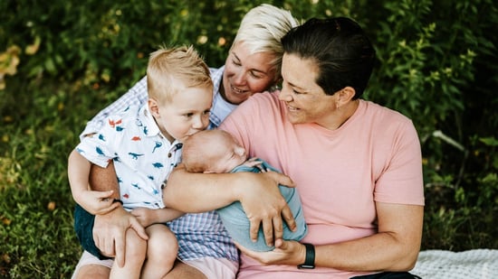 a smiling LGBTQ couple holds their toddler and newborn son against a backdrop of trees