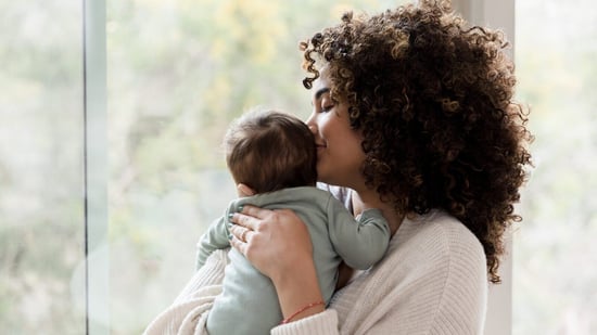 mixed black mother with curly hair holding new baby by window kissing baby's head